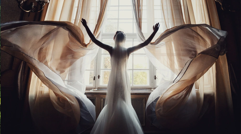 Person in a long white dress stands with arms raised by large windows, curtains billowing around.